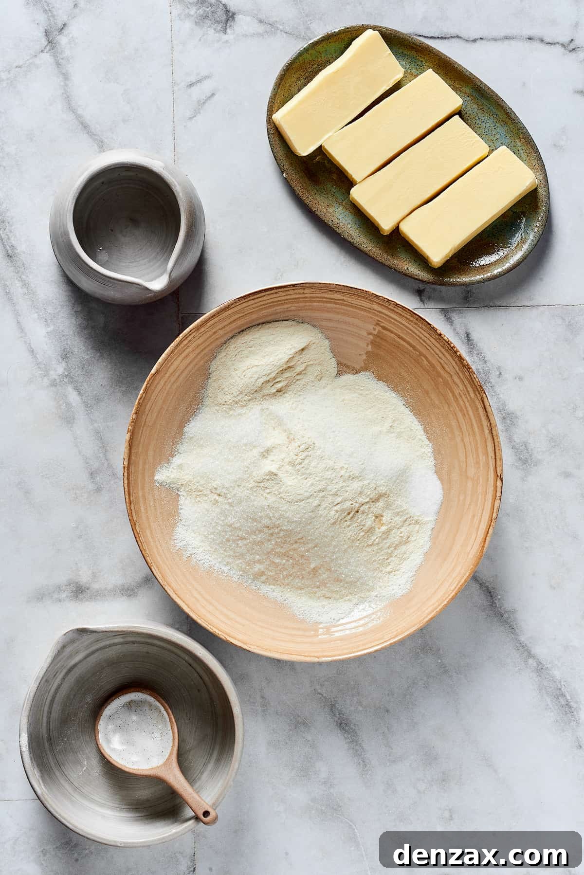 A large mixing bowl containing flour, sugar, and salt, with separate dishes of diced cold butter and a glass of ice water arranged around it.