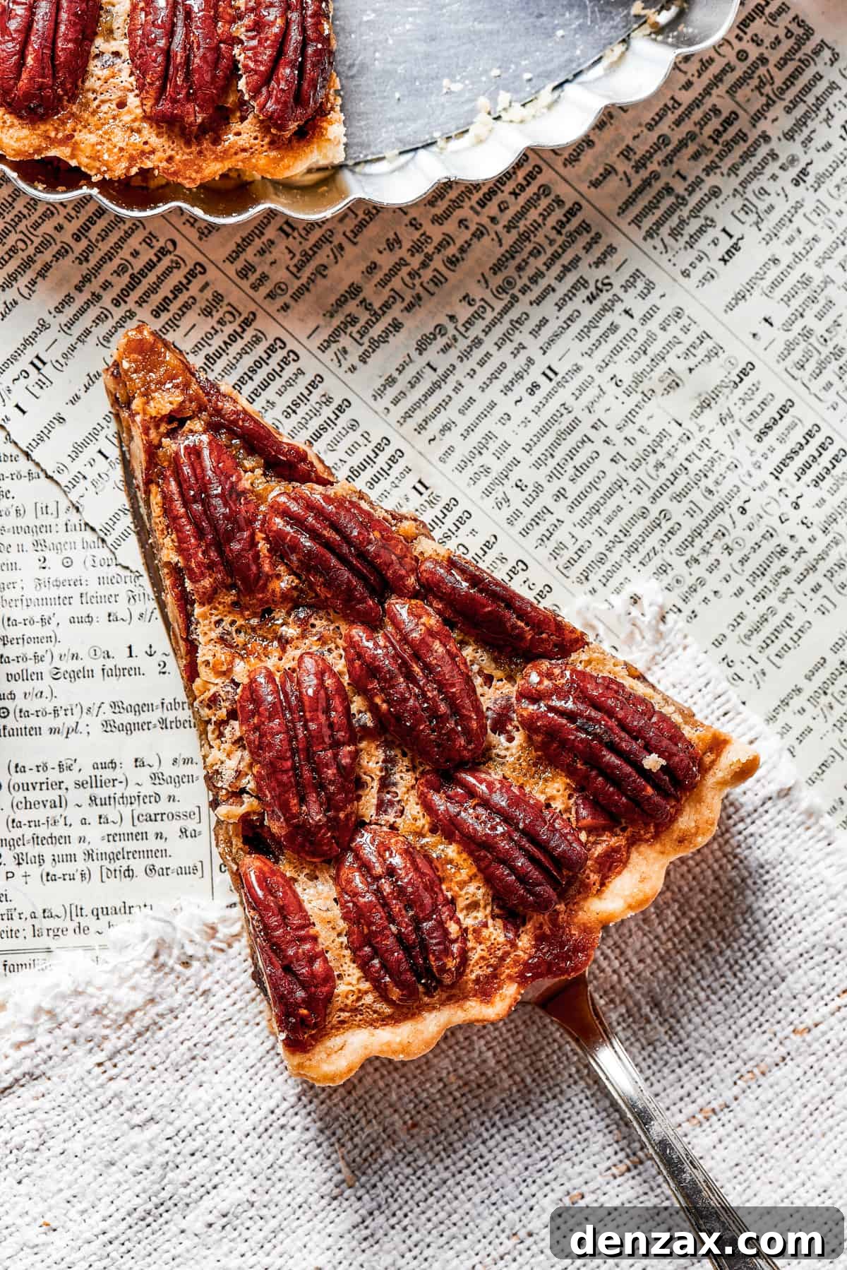 The Best Pecan Pie Recipe is here, easy to make and perfect for your holiday table! You'll love the homemade crust and sweet, nutty filling. An elegant overhead shot featuring a slice of pecan pie held gracefully over a newspaper, with the pie plate positioned above, emphasizing its rustic charm and inviting presentation.