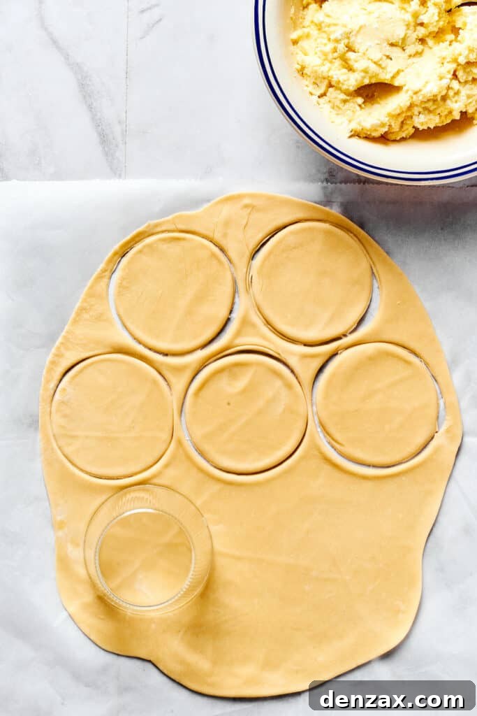 A sheet of thinly rolled dough on a floured work surface, with rounds already cut out. A bowl of cheesy potato filling is nearby.