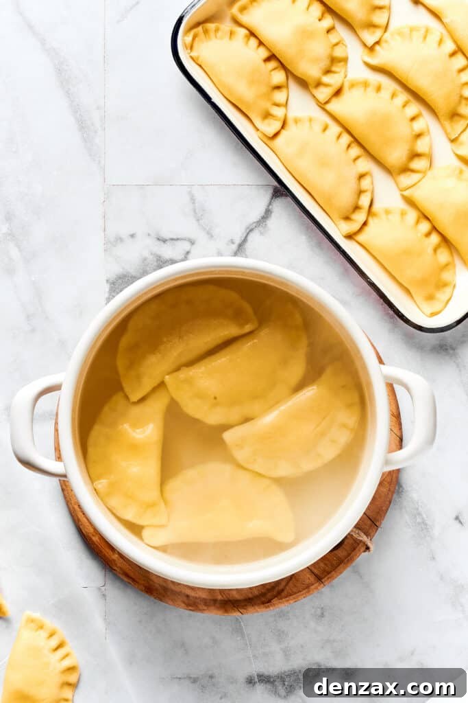 Pierogies boiling gently in a pot of water on the stove. A tray of uncooked pierogies sits beside it.