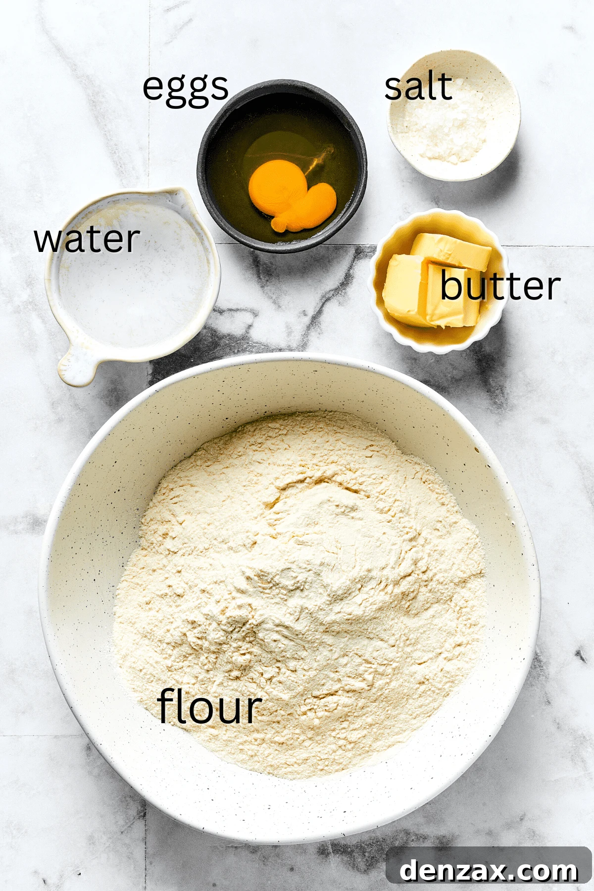 From top: A bowl of water, cracked eggs, salt shaker, butter sticks, and a bag of flour on a wooden surface.