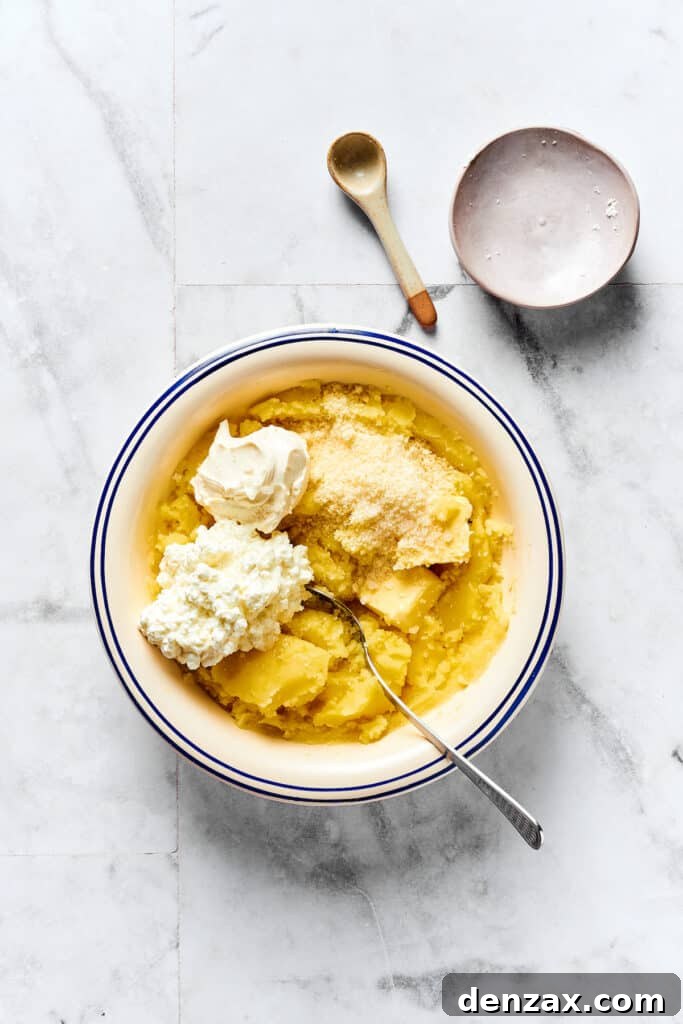 A hand mixing sour cream, cottage cheese, butter, and Parmesan into a bowl of mashed potatoes.