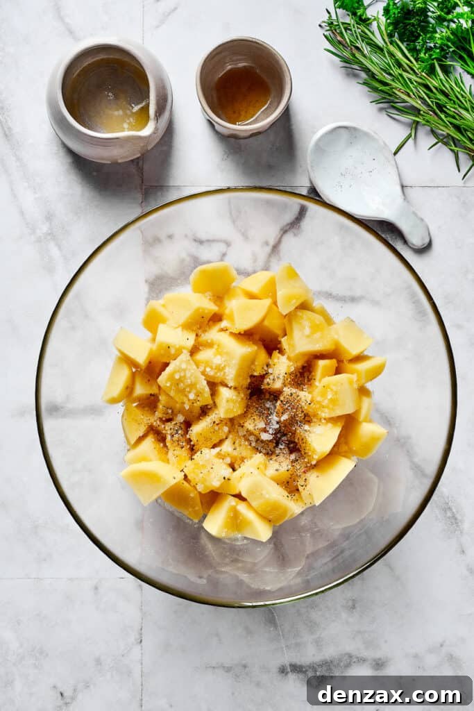Raw potatoes seasoned with melted butter, olive oil, and fresh rosemary in a mixing bowl.
