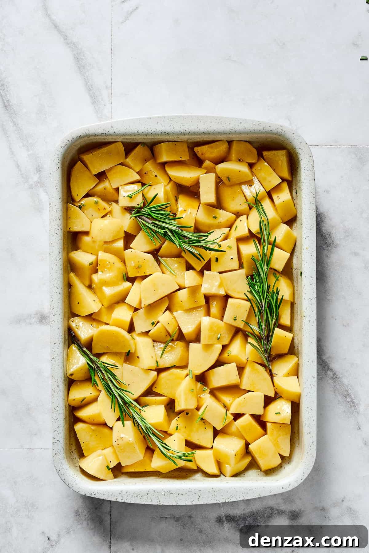 Seasoned raw potatoes and rosemary sprigs spread out evenly in a baking dish, ready for roasting.