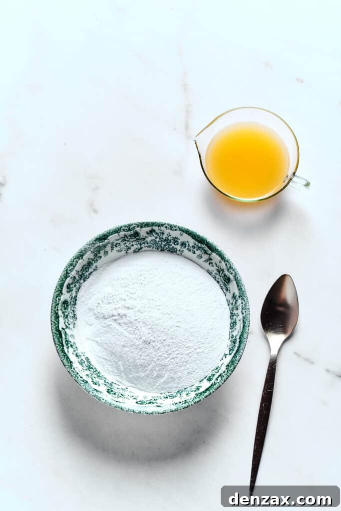 A bowl of powdered sugar next to a glass of fresh orange juice and a spoon, ready for making glaze.