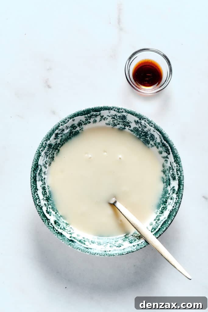 A bowl of creamy, white orange glaze with a small dish of vanilla bean paste nearby, a spoon resting in the glaze.