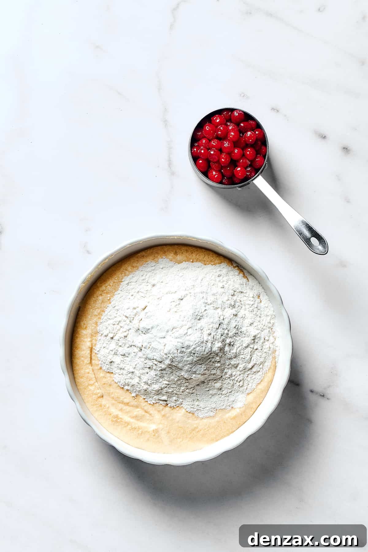 Dry flour mixture being added to the wet ingredients in a mixing bowl, with a bowl of fresh cranberries ready to be folded in.