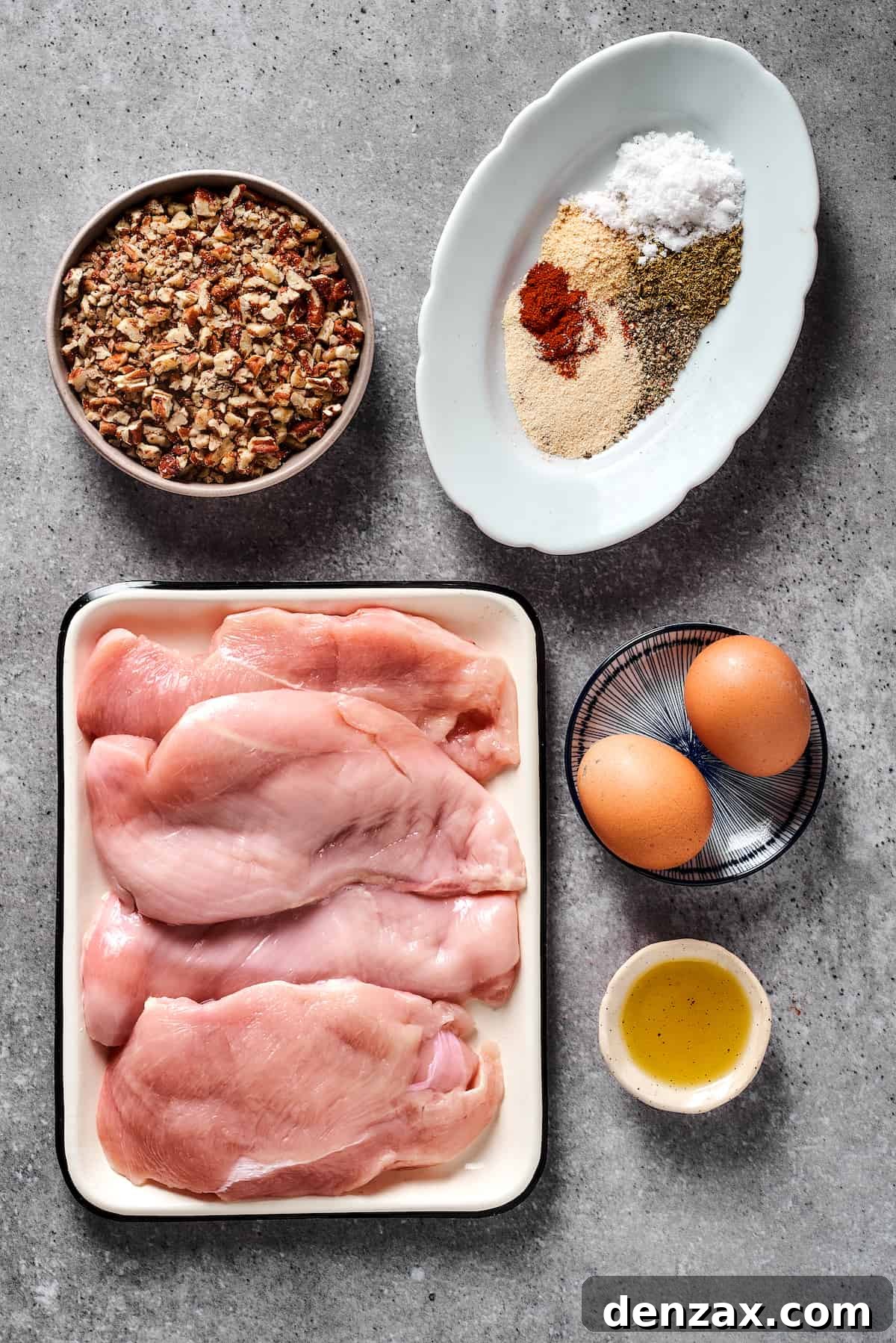 A beautiful arrangement of the key ingredients for Pecan Crusted Chicken: a pile of chopped pecans, a small bowl of mixed spices, a raw chicken breast, beaten eggs in a bowl, and a bottle of olive oil.