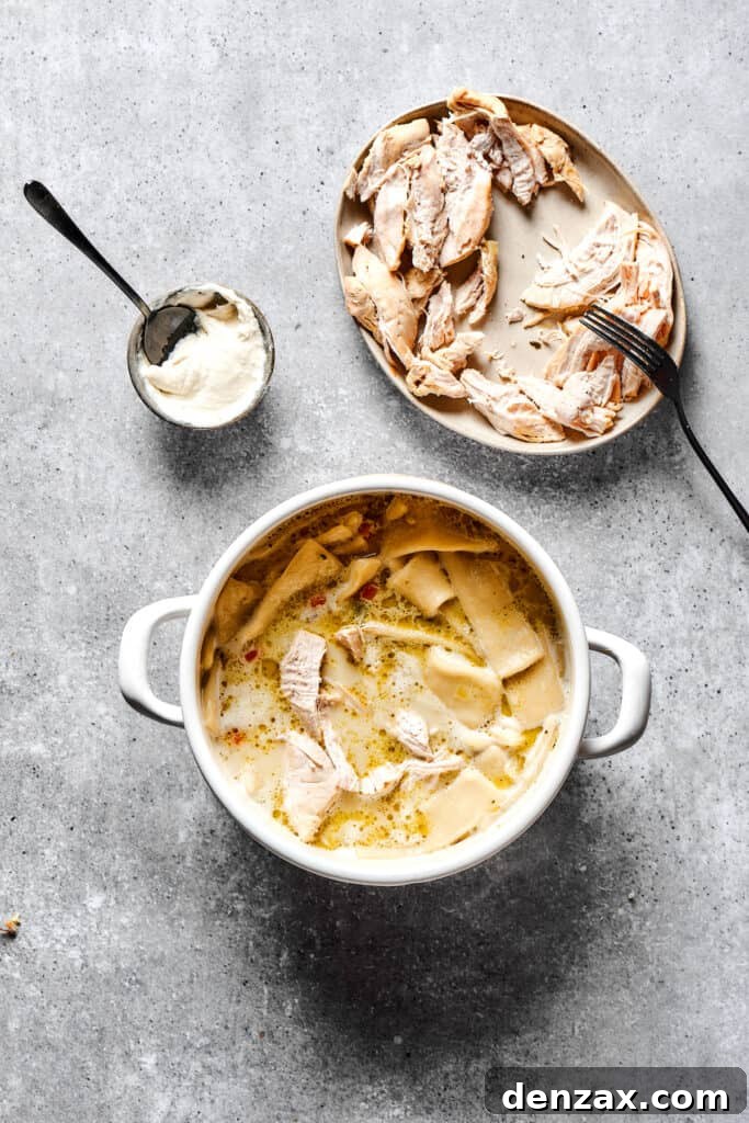 Flat dumplings gently cooking in a creamy soup, with shredded chicken and a measuring cup of thick cream nearby.
