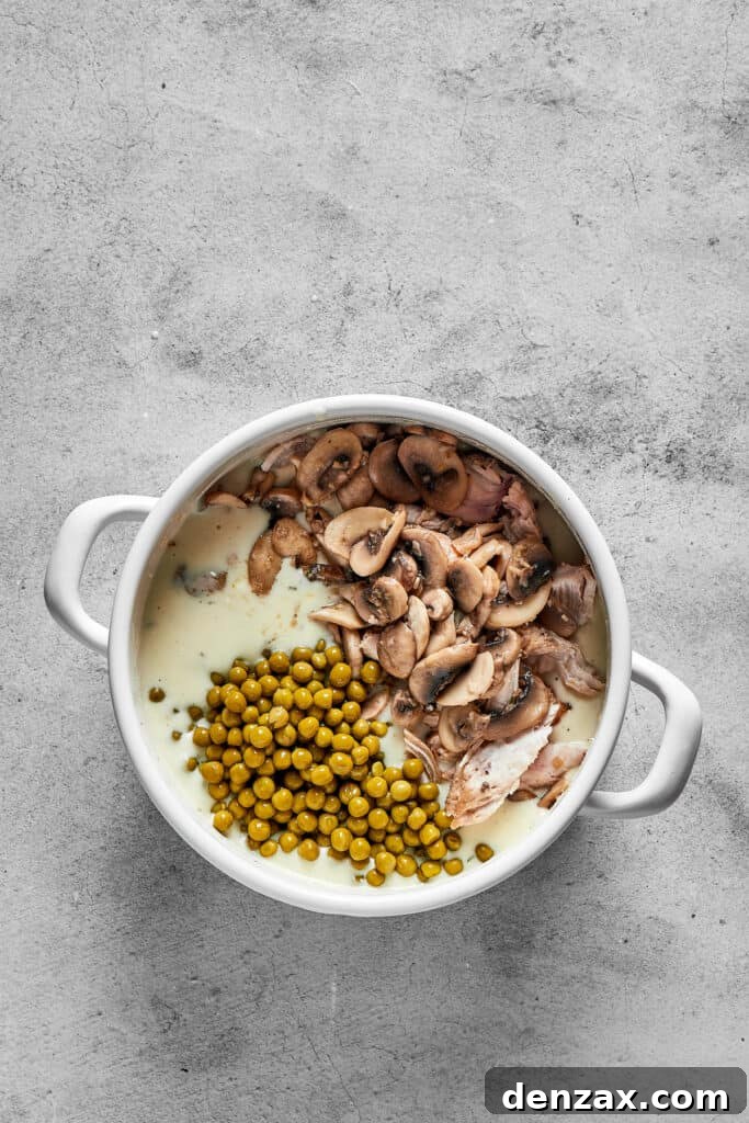 Cooked chicken, mushrooms, peas, and pasta being mixed into the creamy sauce in a large pot.