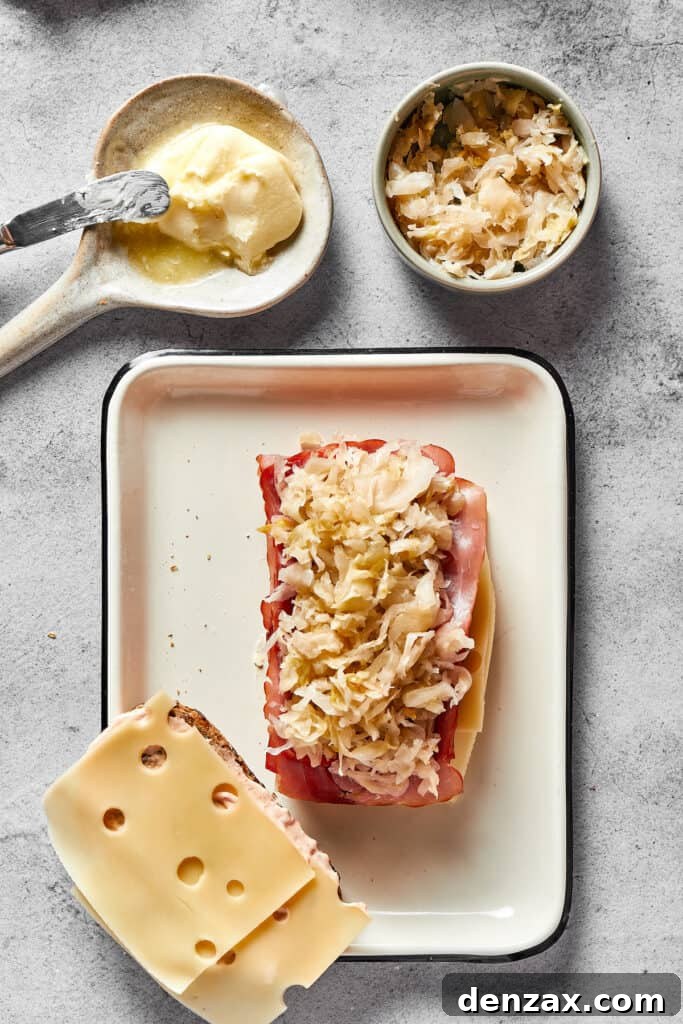 One slice of rye bread is stacked with two slices of Swiss cheese, a generous portion of corned beef, and tangy sauerkraut, positioned next to another slice of rye bread topped only with Swiss cheese, ready for assembly.
