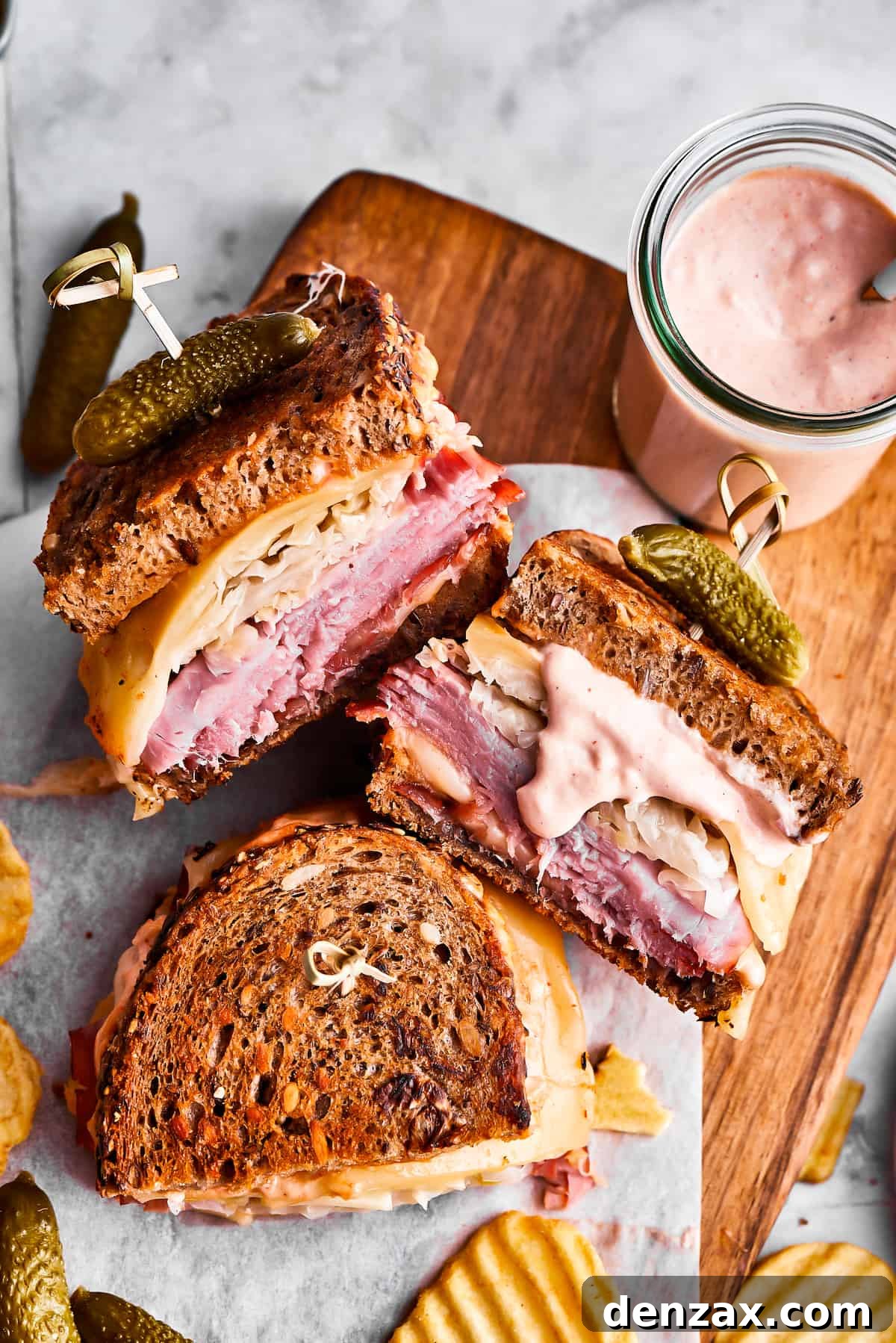 Three perfectly cut Reuben sandwich halves are presented alongside a serving of crispy potato chips and a small jar of Russian dressing on a rustic wooden cutting board.