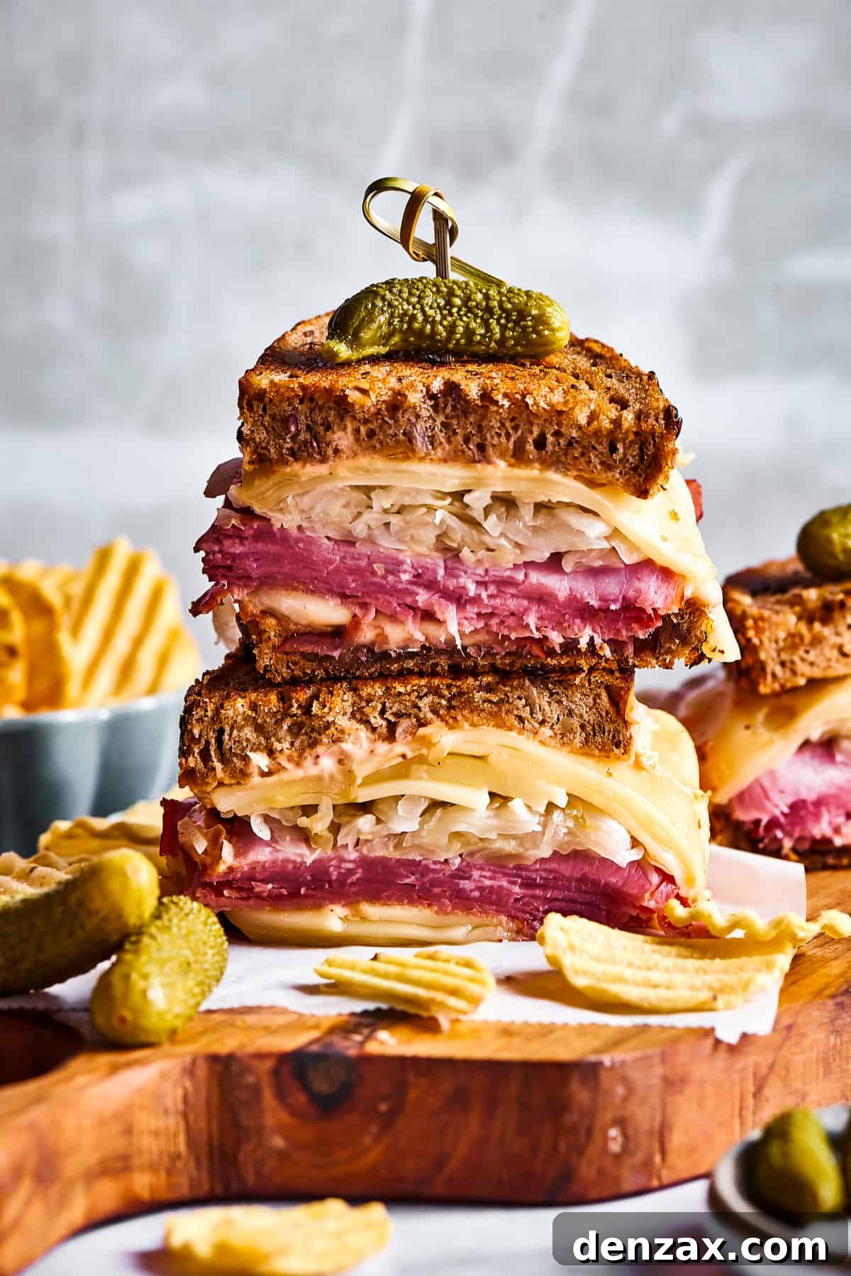 A delectable spread featuring grilled Reuben sandwich halves, a side of creamy Russian dressing, crispy potato chips, and crunchy dill pickles, all artfully arranged on a wooden cutting board.