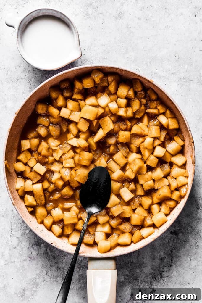 Diced apples gently cooking in a skillet, coated in a sweet, spiced sauce, with a stirring spoon nearby.
