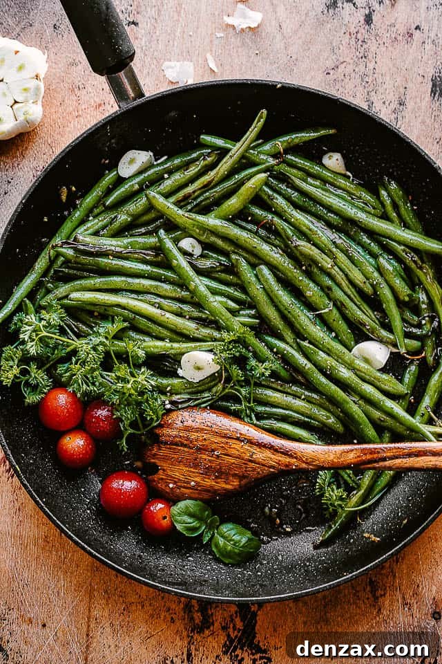 Green beans, cherry tomatoes, and garlic cloves in a black skillet with a wooden spoon, ready for serving