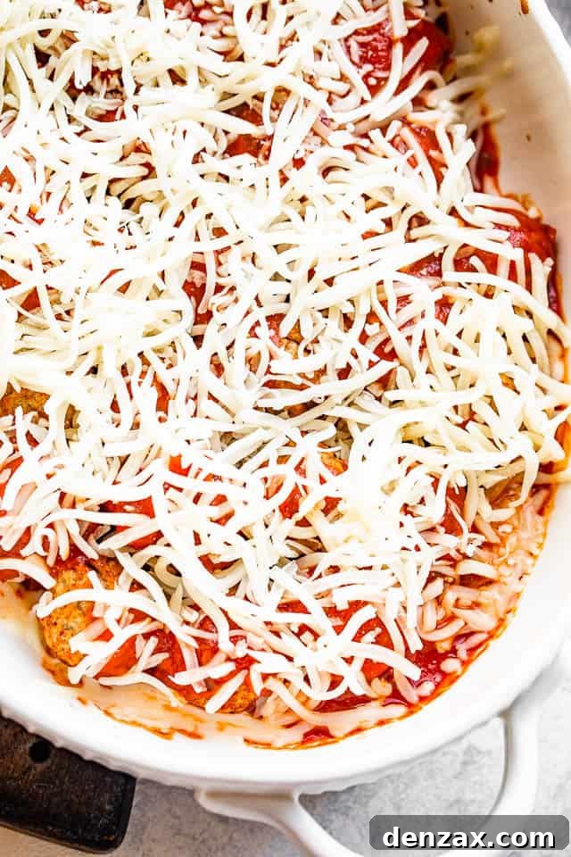 A white baking dish filled with savory meatballs, now coated in rich marinara sauce and sprinkled generously with shredded mozzarella cheese, glistening and ready for its final bake to achieve a perfect melt.