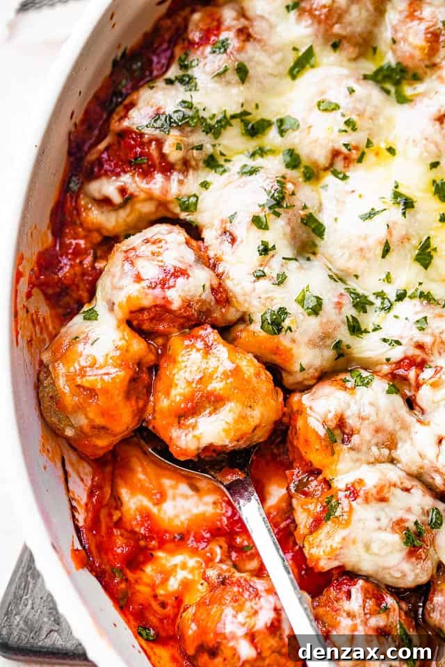 A serving spoon scoops out a portion of the Cheesy Baked Meatball Casserole from a white baking dish, revealing succulent meatballs, rich marinara, and melted cheese underneath.