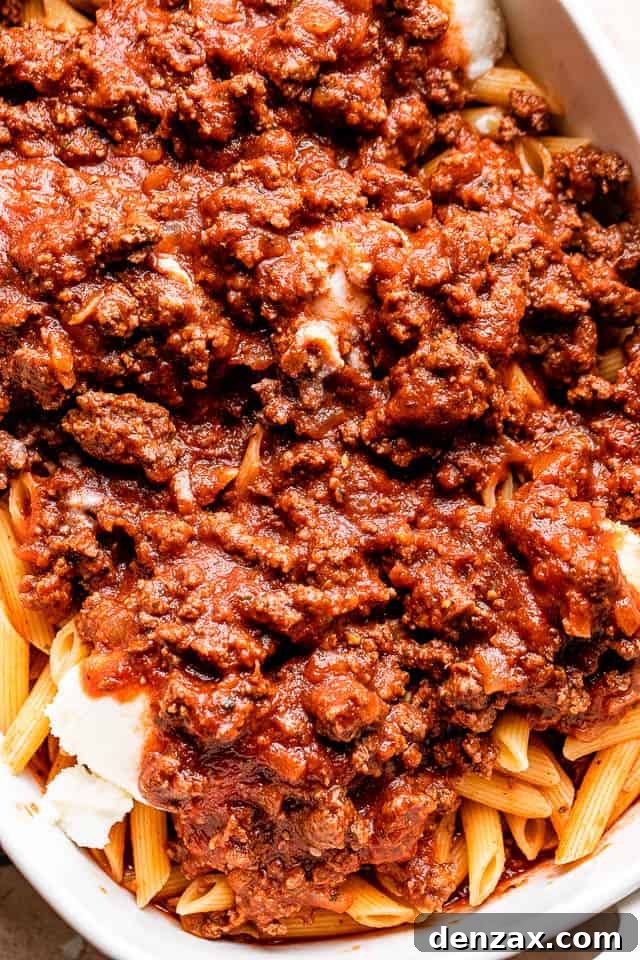 Ground beef and tomato sauce generously poured over perfectly cooked penne pasta in a mixing bowl, ready for assembly.