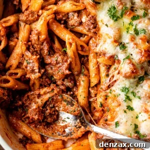 A rich baked ziti casserole, featuring golden-brown melted cheese, nestled in a white baking dish. Tendrils of rich tomato sauce and pasta peek through the cheesy topping, inviting a hearty serving.