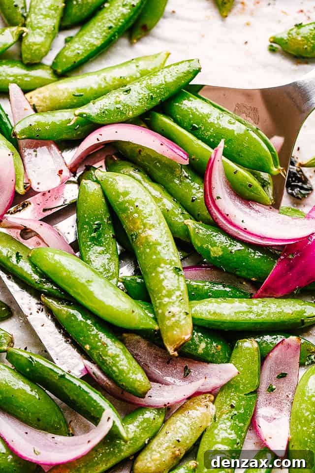 Scooping up perfectly roasted sugar snap peas and red onions from a baking sheet, ready to serve as a delicious and healthy side dish.