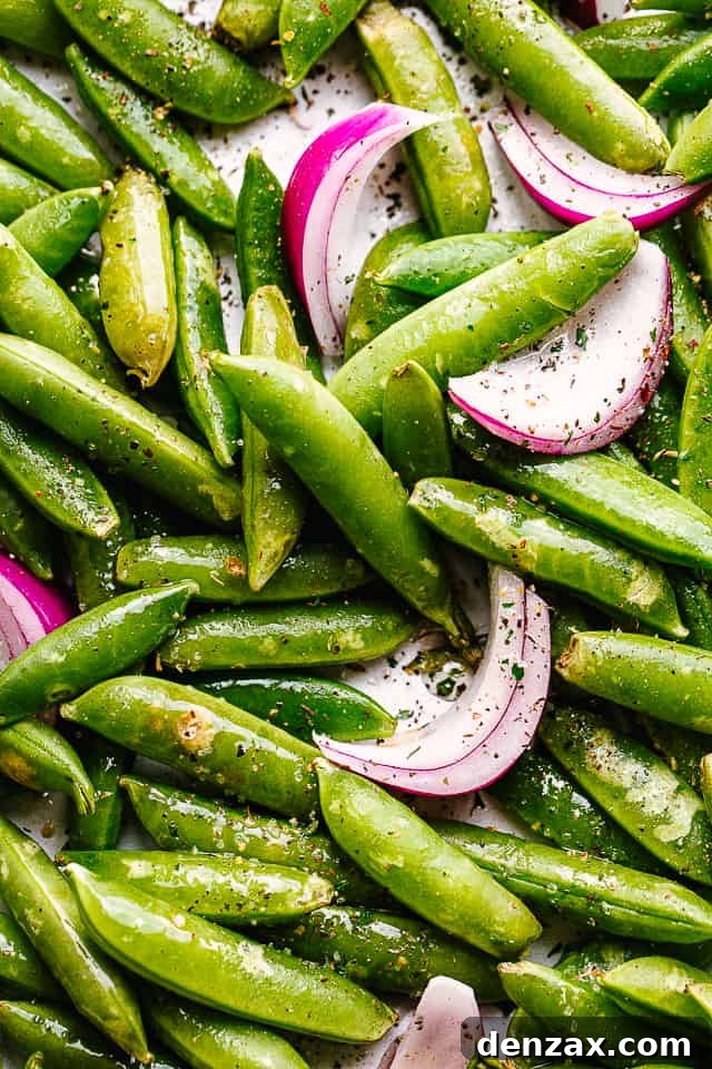 Close-up of fresh, raw sugar snap peas and thinly sliced red onions tossed with olive oil, garlic powder, and Italian seasoning on a baking sheet, ready for roasting.