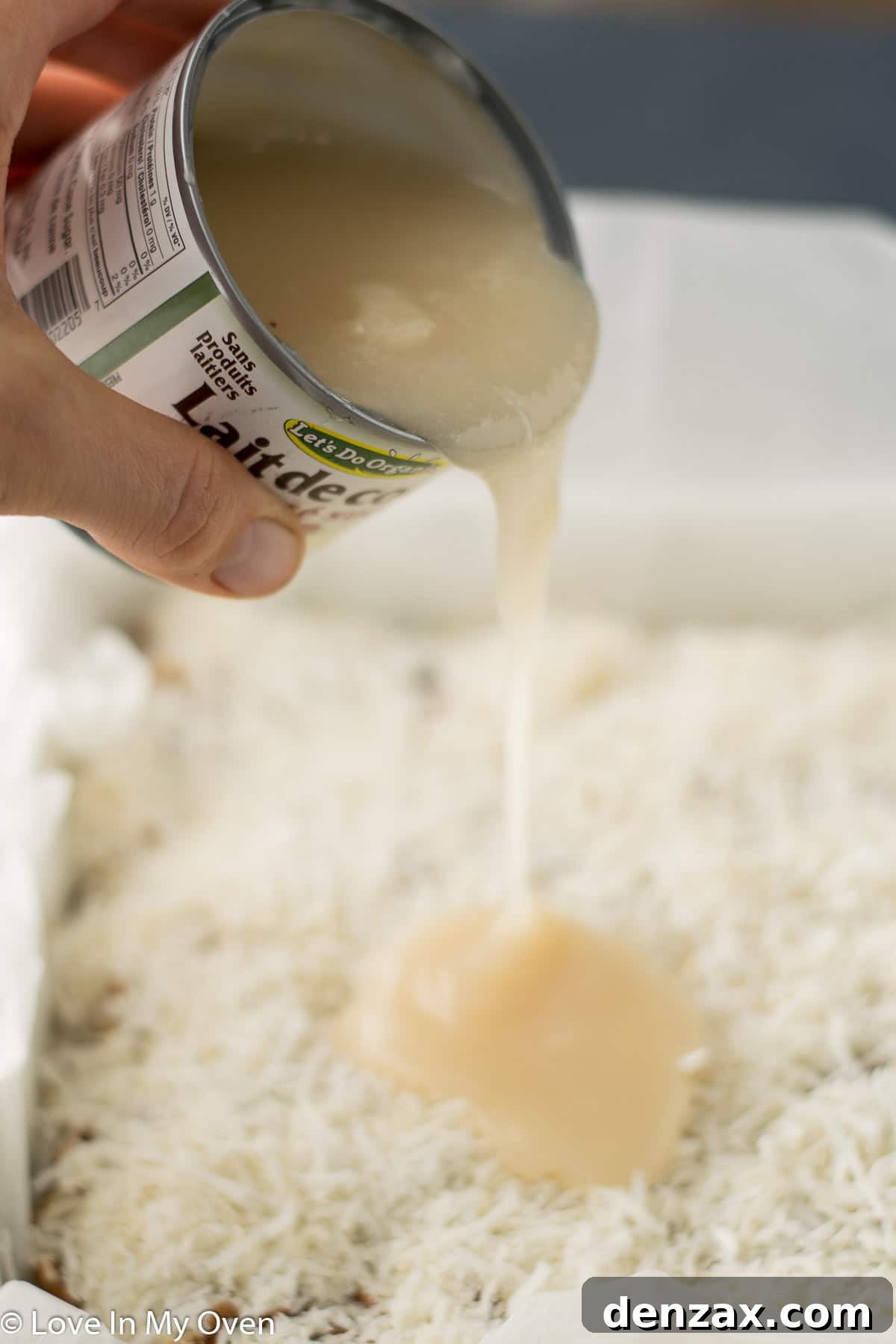 Sweetened condensed coconut milk being poured generously over the layers of vegan magic bars before baking.