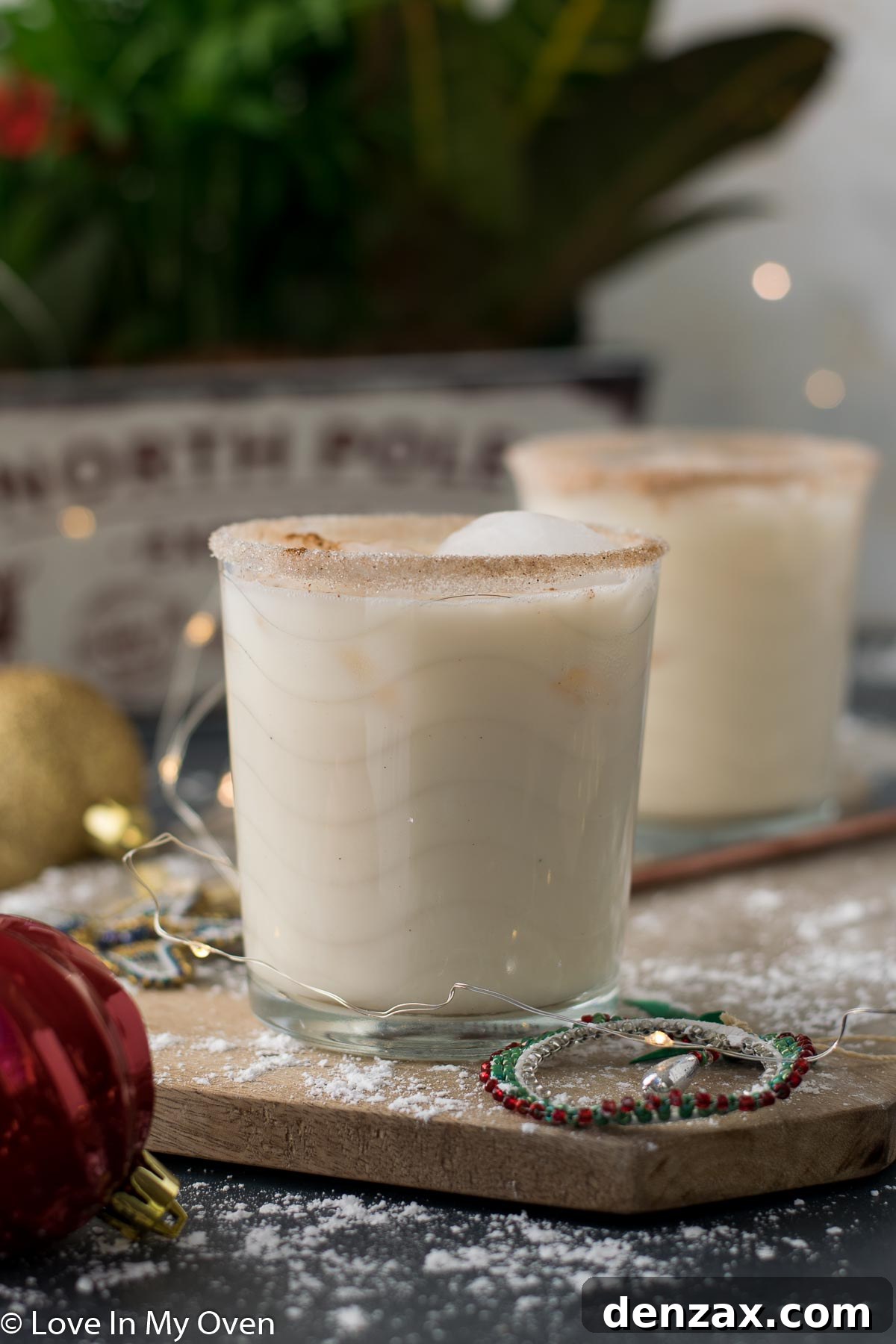 Two festive Vodka Eggnog Cocktails with holiday decorations and lights in the background