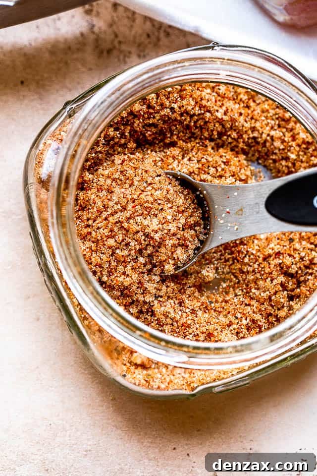 A silver tablespoon rests inside a glass jar filled with a rich, dark brown pork seasoning rub, illustrating the key to flavorful pork chops.