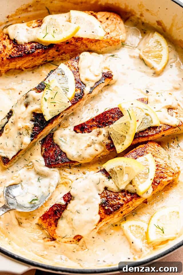 Four salmon fillets topped with creamy dill sauce and lemon slices in a white pan