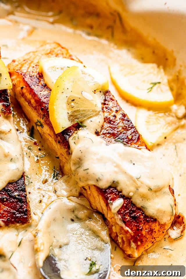 Close up of a cooked salmon fillet generously topped with creamy dill sauce and a slice of lemon