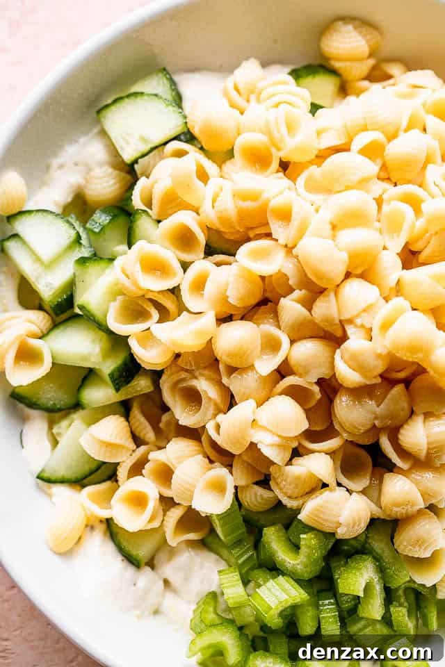Small pasta shells beautifully set over a bed of diced cucumbers and thinly sliced celery in a large white mixing bowl, ready for the shrimp and dressing