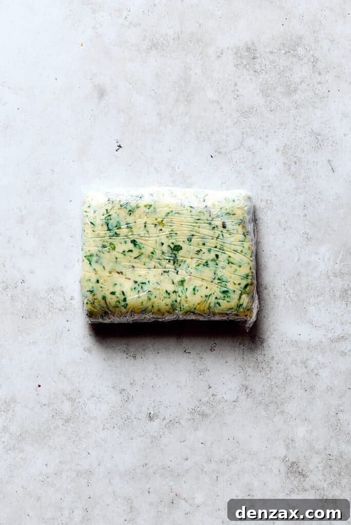 A log of frozen garlic herb butter wrapped in plastic wrap, ready for slicing.