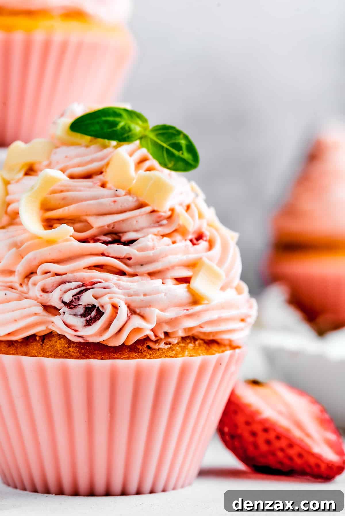 A close-up of a beautifully decorated strawberry cupcake, garnished with fresh mint and a strawberry slice, sitting on a pristine white plate.