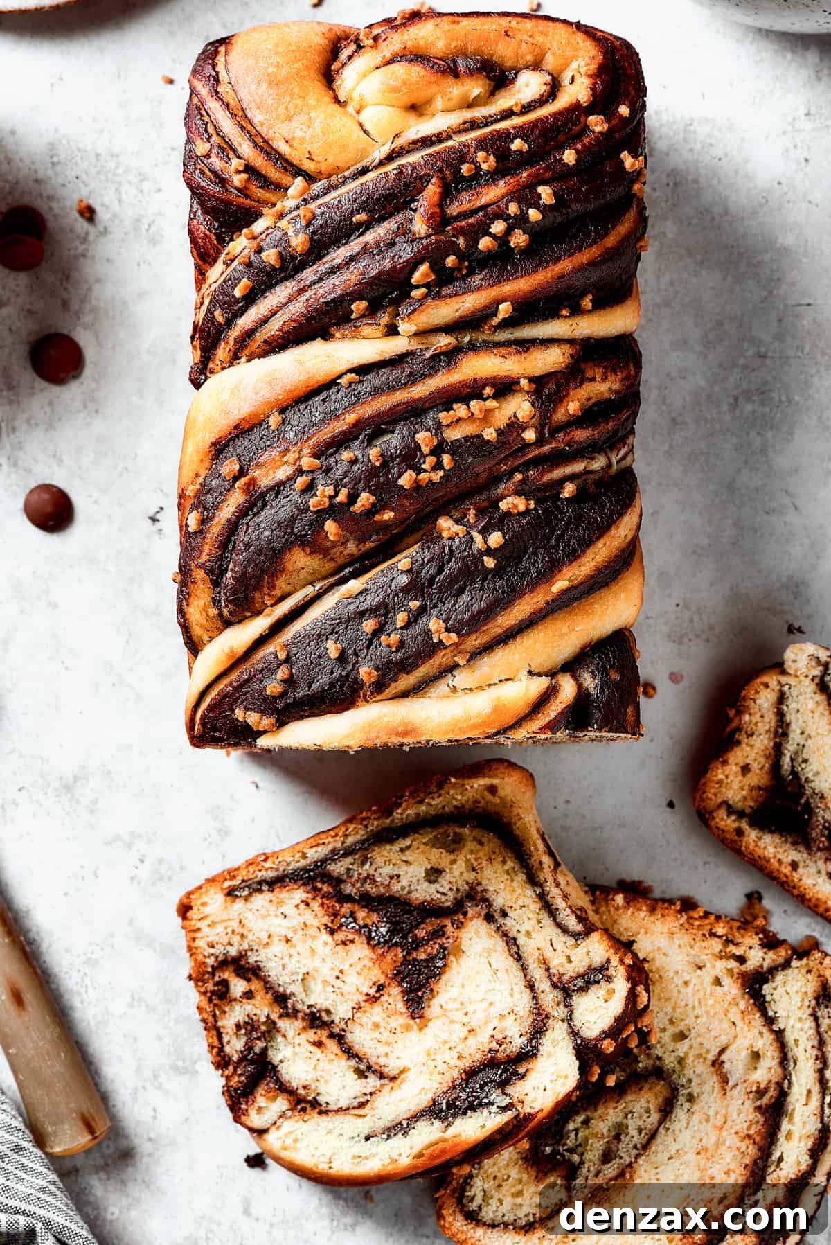 A loaf of chocolate babka that has been partially sliced. The slices are laid out on the work surface, along with a bread knife and a dish of chocolate chips.