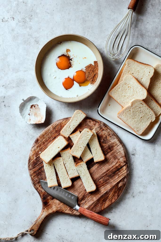 A bowl of eggs, milk, and other custard ingredients next to a tray of sliced bread, and a cutting board of additional bread cut into sticks.