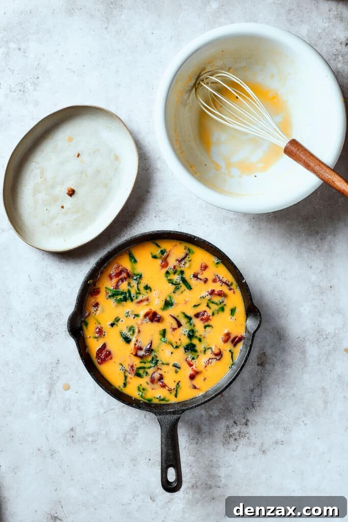 Egg mixture being poured over a bed of cooked bacon, onions, spinach, and potatoes in a cast-iron pan, ready for its stovetop and oven bake.