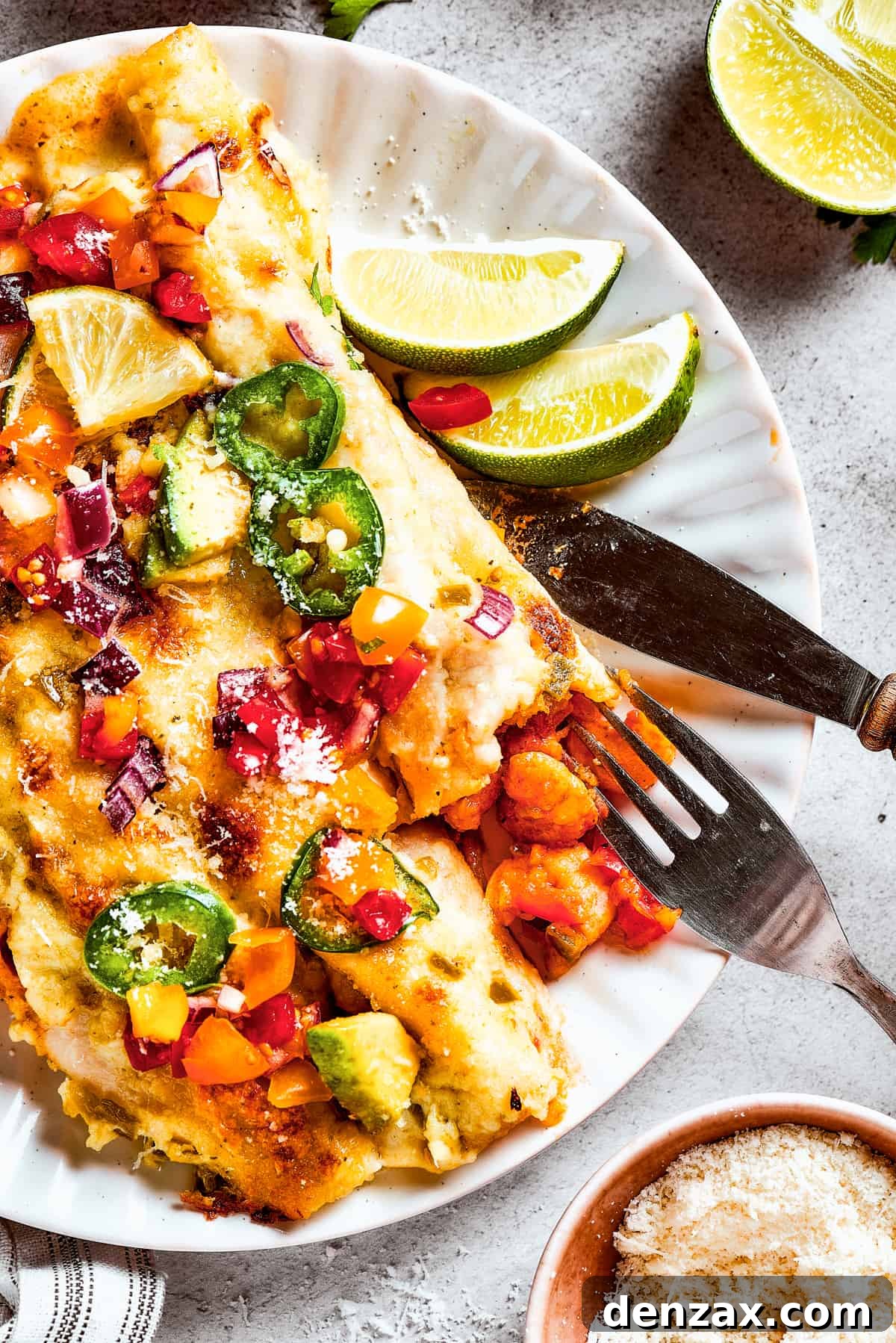 A plate of enchiladas topped with avocado, fresh pico de gallo, and cilantro, served with lime wedges.