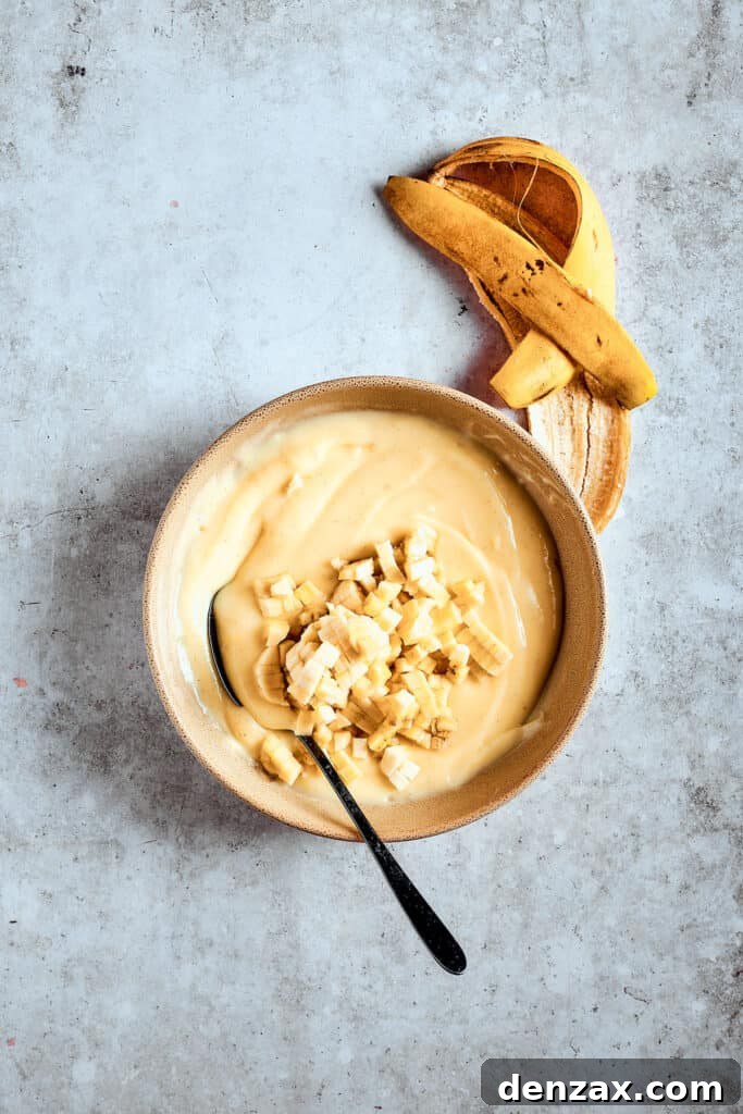Chopped fresh bananas being carefully folded into the cooling banana pudding, ensuring even distribution of fruit.