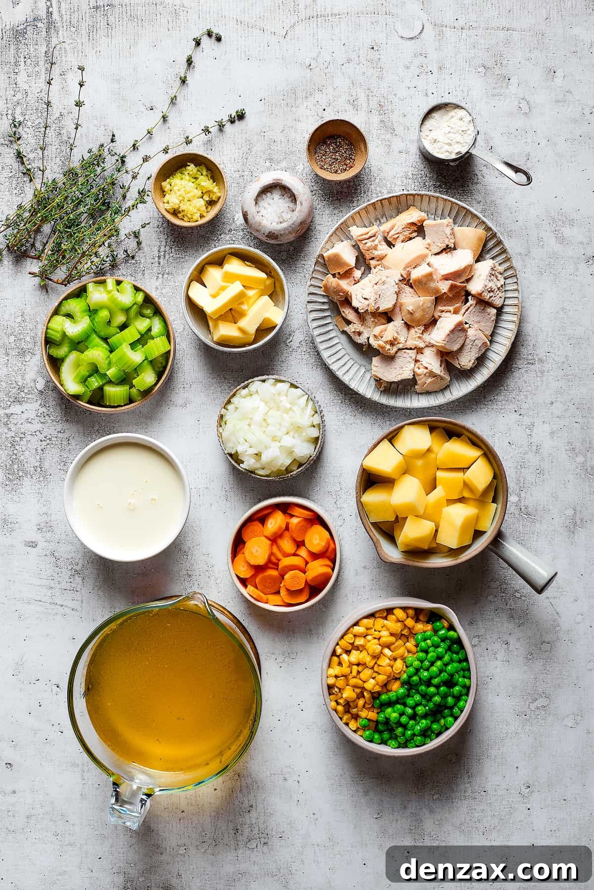 Overhead view of the ingredients needed for chicken pot pie soup: a pyrex of chicken broth, a bowl of cream, a bowl of canned peas and corn, a bowl of carrots, a bowl of potatoes, a bowl of onions, a bowl of garlic, a bowl of celery, a bowl of butter, a plate of cooked chicken, sprigs of thyme, a bowl of salt, a bowl of pepper, and a measuring cup of flour