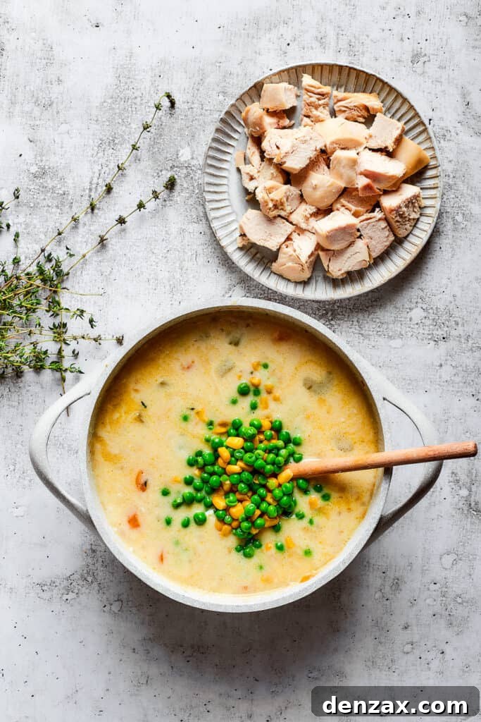 A pile of peas and corn on top of a pot of chicken soup, with a wooden spoon, next to thyme sprigs and a plate of chicken