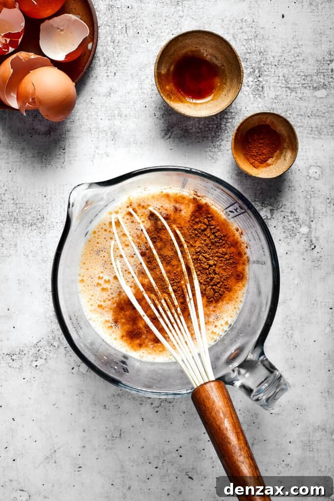 A pyrex filled with eggs and milk beat together with cinnamon on top, with a whisk, next to some bowls and egg shells