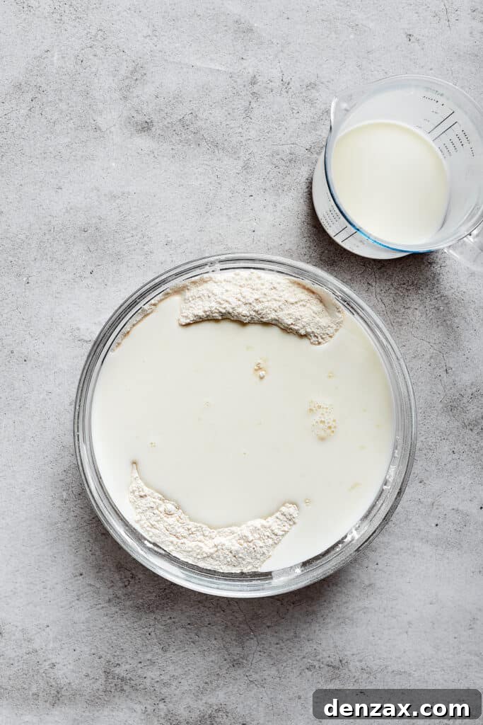 A mixing bowl showing a pool of buttermilk poured over the flour mixture, next to a glass of buttermilk.