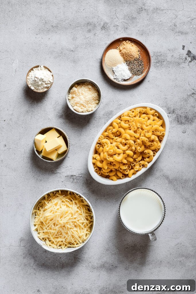 Overhead view of the essential ingredients laid out for making mac and cheese: a bowl of elbow macaroni, a bowl of finely shredded mozzarella cheese, a bowl of freshly grated Parmesan, a glass of whole milk, a small bowl of all-purpose flour, a bowl of unsalted butter cut into cubes, and a separate bowl containing salt, black pepper, garlic powder, and onion powder.