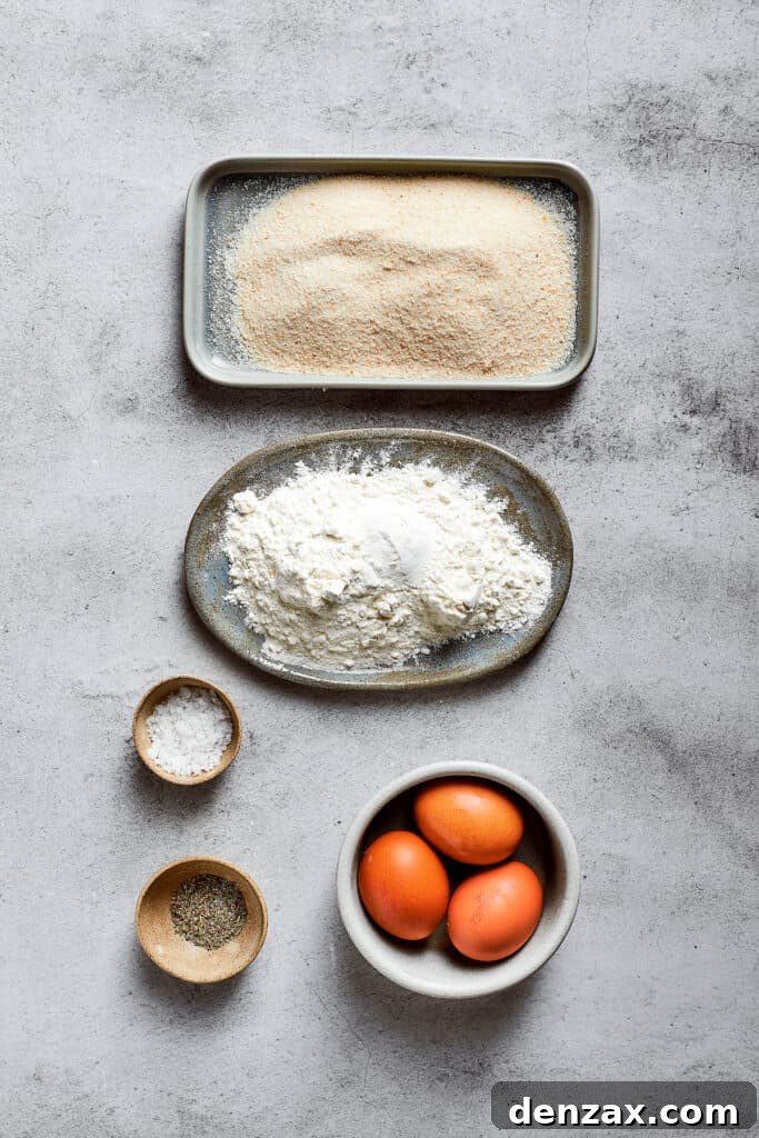 Overhead view of the ingredients prepared for breading the mac and cheese bites: a plate of all-purpose flour, a plate of light and crispy panko breadcrumbs, a small bowl of salt, a small bowl of fragrant Italian seasoning, and three whisked eggs in a bowl.