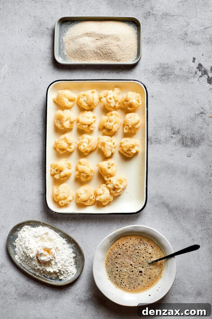 Overhead view of a tray filled with uniformly rolled balls of mac and cheese, neatly arranged next to separate bowls of flour, panko breadcrumbs, and a bowl of beaten eggs seasoned with Italian seasoning.