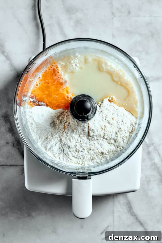 Overhead view of flour, egg yolk, milk, and yeast mixture in a food processor, ready to be combined.