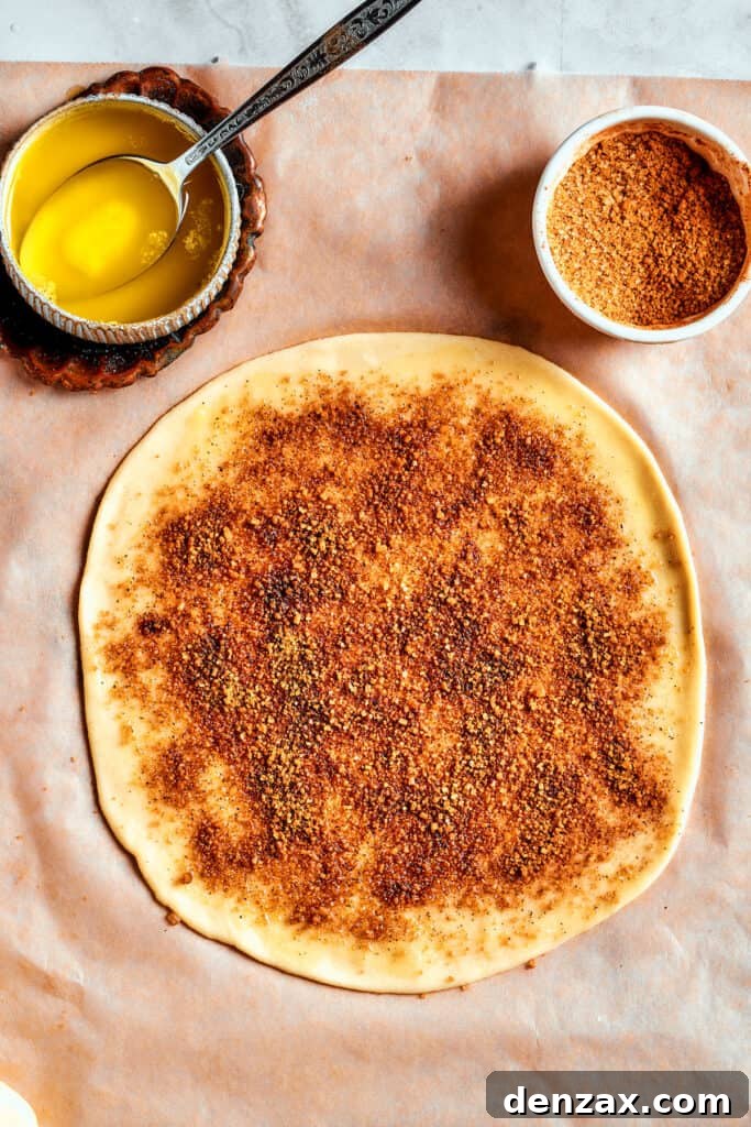 Overhead view of a rolled-out dough circle, already spread with butter and sprinkled with cinnamon-sugar, adjacent to bowls of butter and cinnamon-sugar filling.