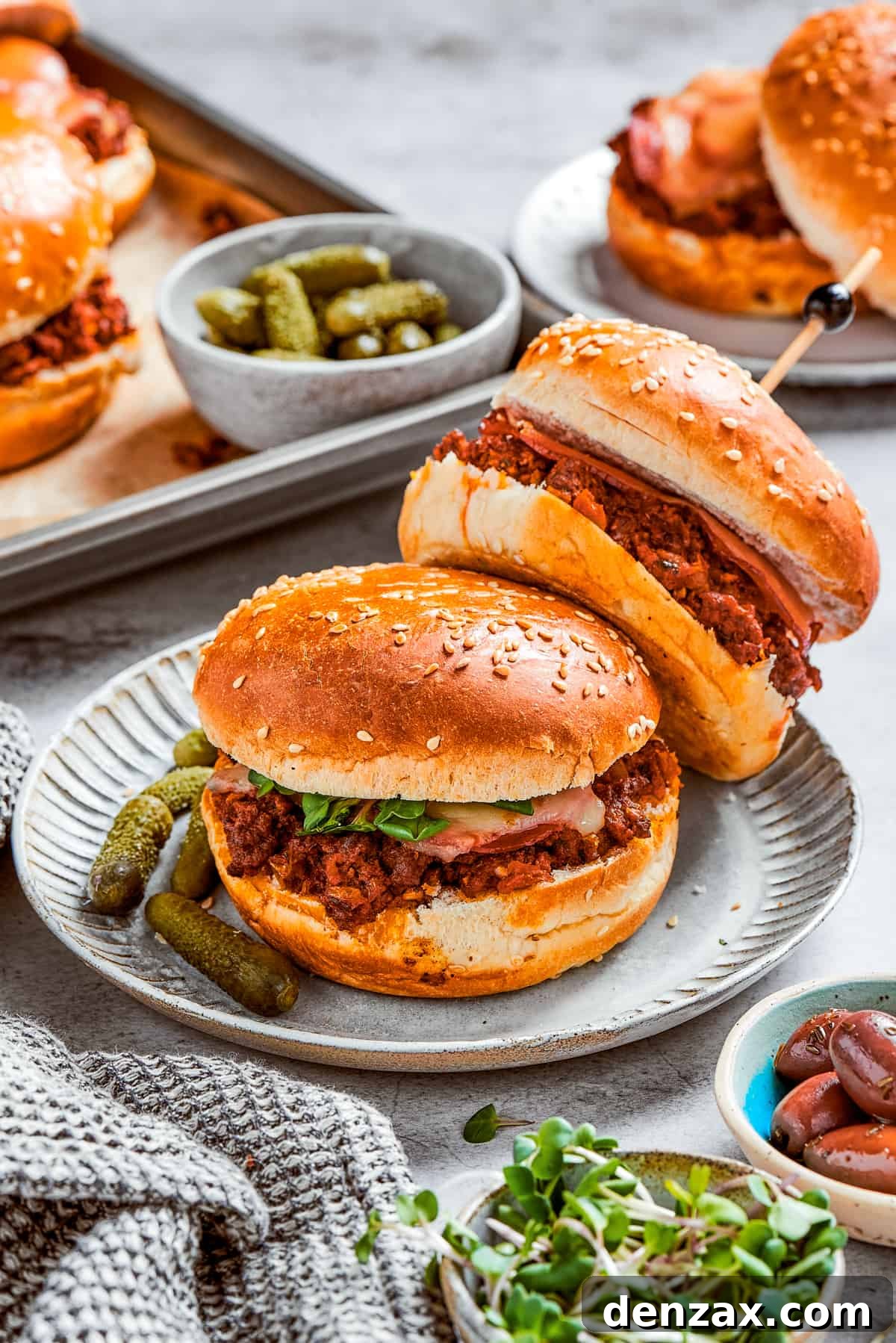 Two perfectly cooked pizza burgers served on a plate, with additional burgers and pickles blurred in the background, showcasing a delicious meal spread.