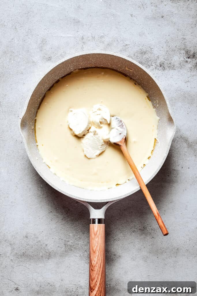 A wooden spoon deftly stirs softened cream cheese into the simmering blend of cream and butter in a white skillet, ensuring a perfectly smooth and luscious Alfredo sauce is created.