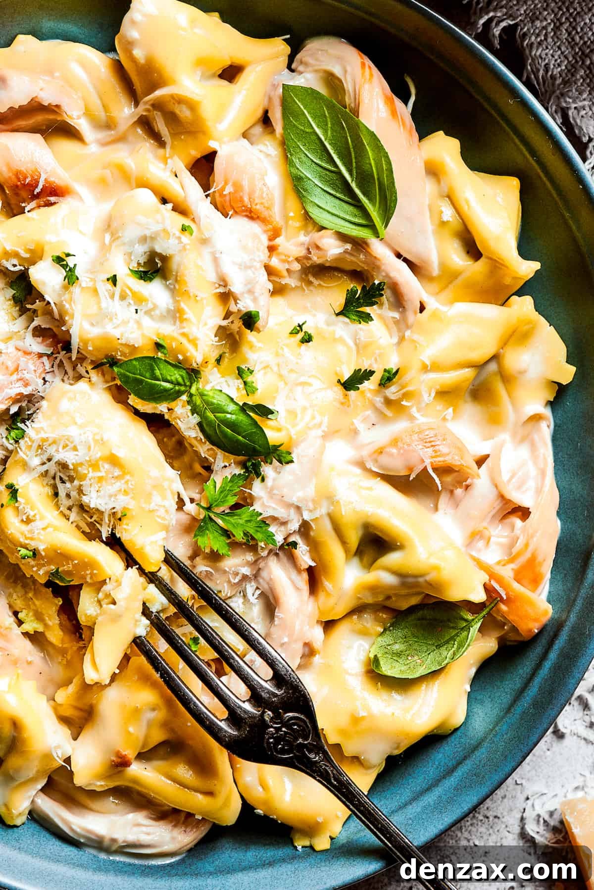 A vibrant blue bowl filled with a generous portion of creamy Chicken Tortellini Alfredo, artfully garnished with a sprinkle of freshly grated Parmesan cheese and vibrant fresh basil leaves, ready to be enjoyed as a delightful meal.